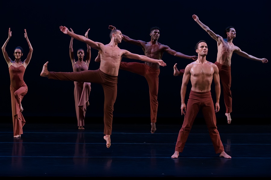 two women of the chorus balance on one leg with their arms extending upward while a trio of men jump into the air with their right leg extended to the side. to the front of that action a man makes a wide stance his torso perfectly straight and his eyes gazing upward in thought.